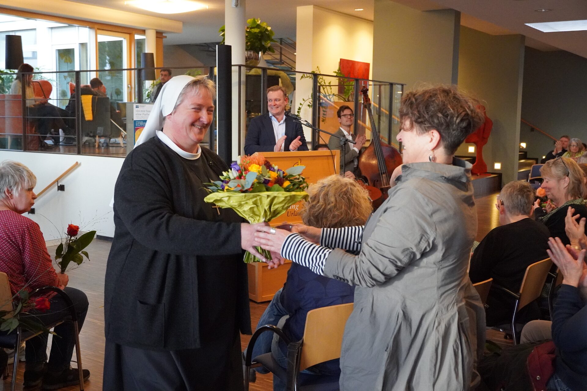 In einem bestuhlten großen Raum bekommt eine Frau einen Blumenstrauß überreicht, während drum herum die Zuschauer klatschen